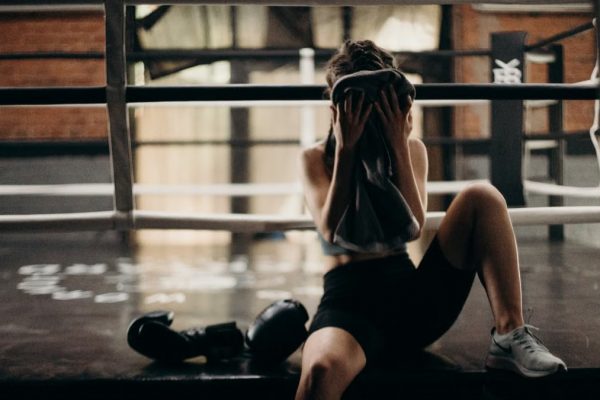 A female boxer sitting in a gym ring, resting and wiping sweat with a towel after a workout.
