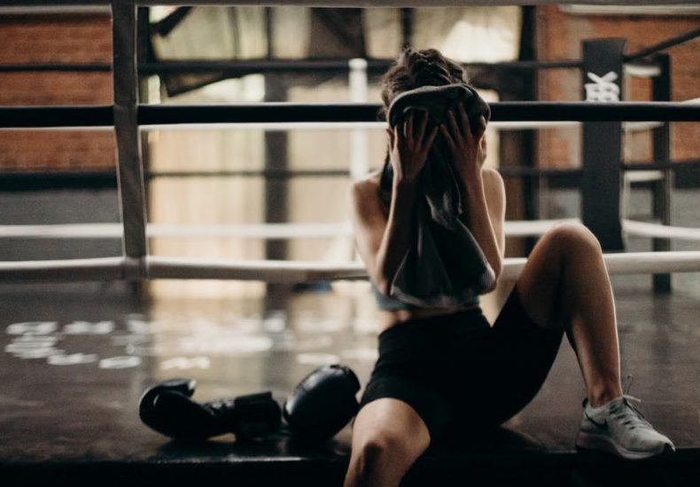 A female boxer sitting in a gym ring, resting and wiping sweat with a towel after a workout.